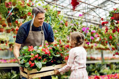 Happy father carrying a crate with flowers while his small daughter is helping him at garden center. 