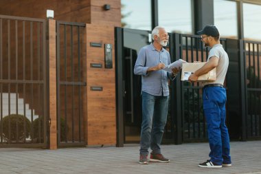 Happy mature man signing paperwork while communicating with delivery man in front of his home. 