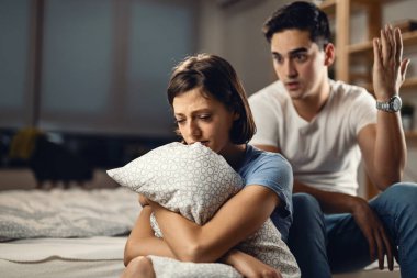 Young couple having relationship difficulties and arguing in the bedroom. Focus is on sad woman holding a pillow. 