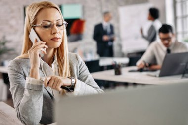 Pensive businesswoman talking to someone over mobile phone while working at her office desk.  There are people in the background. 