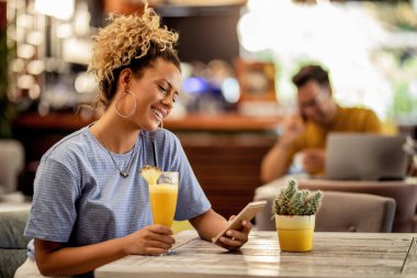 Smiling woman reading text message on cell phone while sitting in a cafe and drinking juice. 