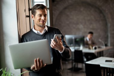 Happy businessman standing by the window in the office and reading text message on mobile phone while holding laptop. 