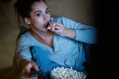 Young woman feeling shocked while watching movie and eating popcorn in the evening at home. 