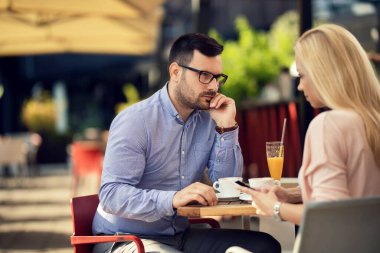 Bored man looking with suspicion at his girlfriend who is text messaging on cell phone and ignoring him in a cafe,
