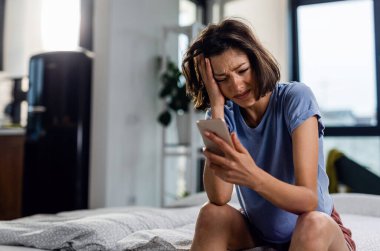 Unhappy woman crying while sitting on the bed and reading text message on her cell phone.