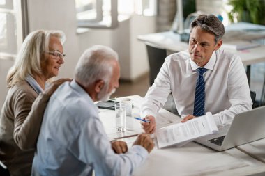 Senior couple analyzing documents before signing them while having a meeting with insurance agent. Focus is on agent. 