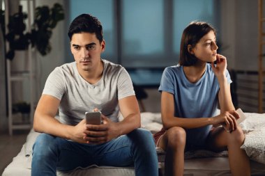 Displeased couple ignoring each other while siting on the bed after the argument.  Focus is on man. 