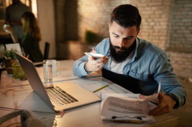 Young businessman working late on paperwork and talking over smart phone's speaker in the office. 