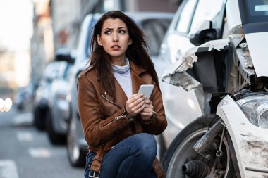 Young unfortunate woman using smart phone and text messaging after a car accident in the city.  