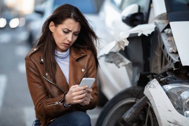Desperate woman typing on mobile phone while being next to her wrecked car after the accident. 