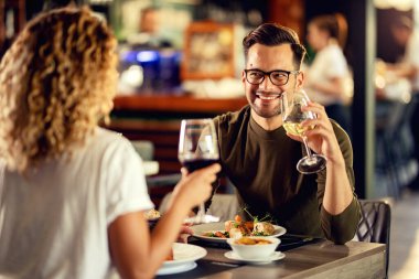 Young happy man drinking wine and talking to his girlfriend while having lunch in a bar. 