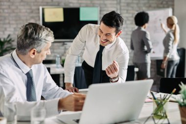 Two businessmen communicating while using computer in the office. Focus is on young happy businessman. 