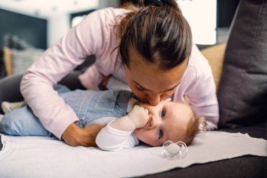 Young loving mother kissing her baby boy on cheek with affection while spending time with him at home. 