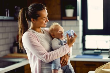 Young caring mother using baby bottle and feeding her baby son in the kitchen. 