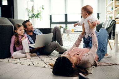 Playful mother holding her baby son while lying on the floor in the living room. Father and daughter are using laptop. 