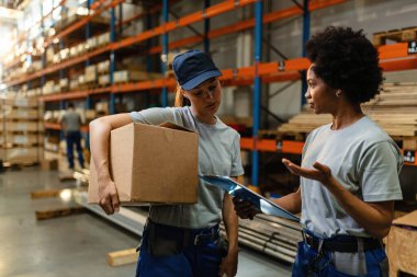 Black warehouse worker and her younger colleague talking while going through shipment list in a storage room. 