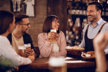 Happy waiter having fun with his customer while serving them with food in a bar. 