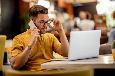 Pensive freelancer talking on cell phone while reading problematic e-mail on laptop in a cafe. 