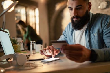 Young businessman using smart phone and credit card while shopping on the Internet at night the office. 