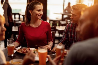 Group of young people having lunch and talking to each other in a pub. Focus is on happy woman. 