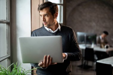 Businessman with laptop standing by the window and day dreaming while looking through it. 