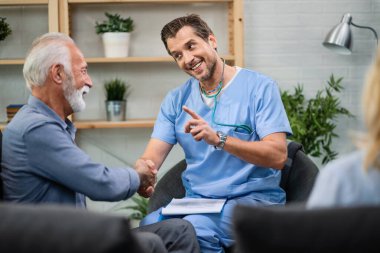 Happy doctor communicating with senior man while shaking hands with him during a home visit. 