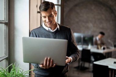 Happy businessman standing by the window and reading an e-mail on a computer in the office. 