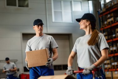 Warehouse worker talking to female colleague while working with carboard boxes in industrial storage compartment. 
