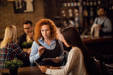 Young waitress with touchpad communicating with female customer who is looking at the menu in a tavern. 