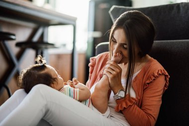 Young loving mother enjoying with her baby daughter and kissing her small feet at home. 