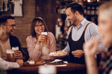 Happy customers drinking beer and having fun with a waiter who is serving them food in a tavern. 