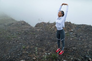 Active sportswoman preparing for morning run and stretching while standing near the cliff on cold misty weather. Copy space.  