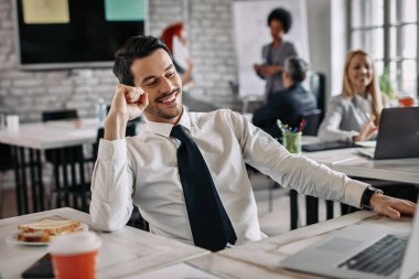 Cheerful businessman celebrating success while working on a computer in the office. There are people in the background. 