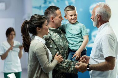 Happy senior doctor talking to little boy who came with mother and military father at clinic. Focus is on military man. 