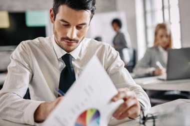 Young businessman analyzing charts and taking notes while working on new project in the office. 