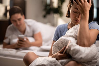Young sad woman with headache sitting in the bedroom while her boyfriend is text messaging on mobile phone in the background.