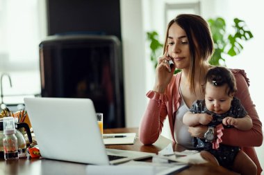 Working mother with baby daughter communicating on mobile phone while reading an e-mail on the computer. 