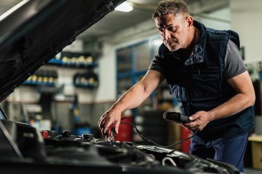Mid adult mechanic using diagnostic tool while checking voltage of a car battery in auto repair shop. 