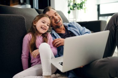 Happy father and daughter having fun while surfing the net on laptop in the living room. 