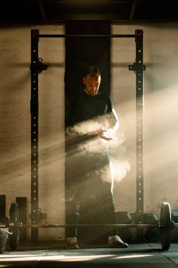 Full length of athletic man clapping hands with talcum powder before lifting  barbell on weight training in a gym.