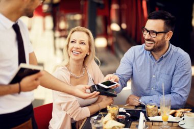 Happy man paying for lunch with smart phone contactless payment while being with girlfriend in a bar. 