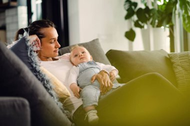 Young mother holding her baby and looking at him while sitting on sofa at home. 