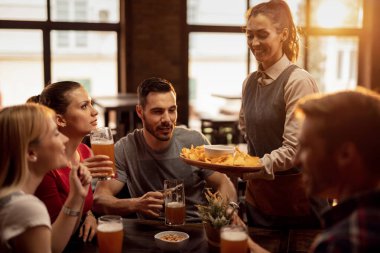 Group of happy friends drinking beer in a tavern while waitress is serving them nacho chips.