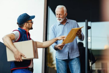 Mature man communicating with a deliverer while standing at front door and receiving a package. 
