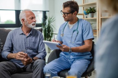 Serious doctor writing medical report while talking with senior man during home visit.  
