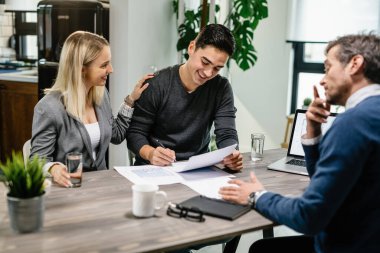 Young happy man signing mortgage contract while being with his wife on a meeting with real estate agent.