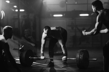 Black and white photo of determined athlete lifting barbell in a gym while his athletic friends are supporting him.