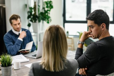 Young man talking to his wife and feeling uncertain while having a meeting with their financial advisor at home.