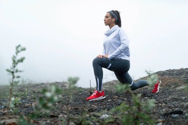 Determined sportswoman stretching in lunge position on a cliff during misty morning. Copy space.
