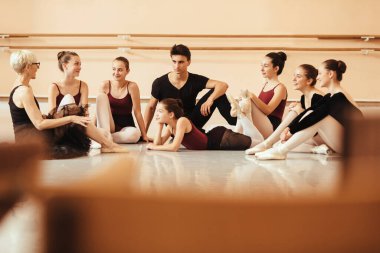 Mature ballet teacher and her students relaxing on the floor and communicating at ballet studio. 
